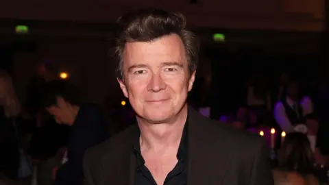 Getty Images Rick Astley smiles at the camera. He is wearing a black shirt, unbuttoned to his chest, and is standing in a dark room at an awards ceremony