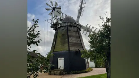 Fulbourn windmill, an early 19th Century building. It has a black base and above its white door is weathered looking boarding which rises to a conical top. Its white sails are on the far side of the mill. Its white fantail is on this side of the mill. It is surrounded by block paving and a house can be glimpsed on the left. 