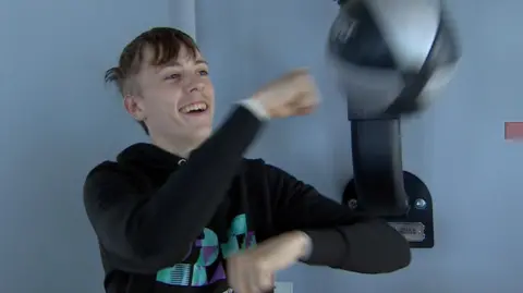 A boy punches a boxing punchbag. He is wearing a black hoody and laughing.