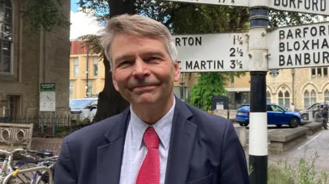 Andrew Gant, a man whose short hair is swept to the left, is pictured wearing a dark suit over a light blue shirt and red tie. He is smiling at the camera. Behind him is a sign that provides the distance to various locations across Oxfordshire. 