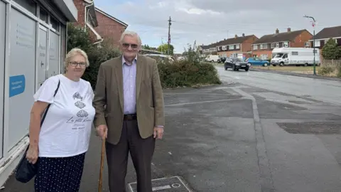 A woman and a man standing on a pavement. The woman has short blonde hair, a white T-shirt and black trousers with white spots. The man has white hair, black tinted glasses. He is wearing a brown/green suit jacker, blue shirt and black trousers and is holding a walking stick.