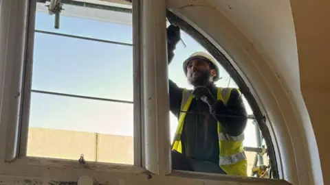 North Northamptonshire Council A man, working on a window, wearing a white hard hat, black top, gloves and high-viz jacket. He has one arm up and scaffolding behind him. 