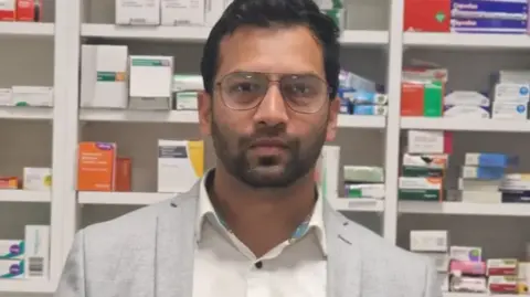 A man with a very short beard is wearing glasses, a white shirt and a grey blazer. He is standing in front of row of medication boxes.