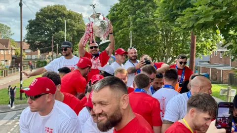 A view from the top of an open-top bus as it travels along a tree lined street. A number of people are taking selfies and waving to crowds on the pavement.