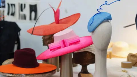 A collection of four hats on mannequins - a the back is an orange and pink hat with orange feather, in front of that is a orange and raspberry colour hat, a bright pink and light pink hat in the middle with a bow and then a blue hat to the far right. 