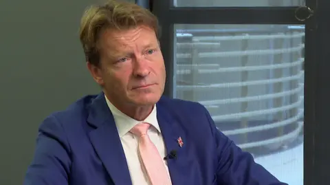 BBC Richard Tice wearing a blue suit, white shirt and cream coloured tie as he sits looking towards the interviewer who is out of shot. He has a Union jack pin badge on his lapel.