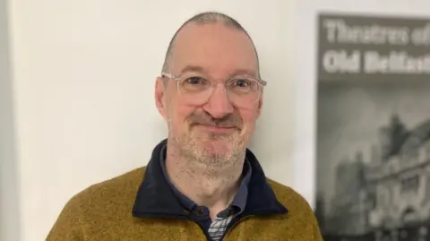 Stuart Marshall is wearing a yellow fleece and round spectacles. He is stood next to a poster which says 'Theatres of old Belfast