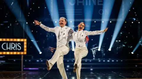 Chris McCausland and Dianne Buswell wearing white suits during the live show on Saturday, with their arms spread out and one leg up, both holding each other, and animated towards the camera.