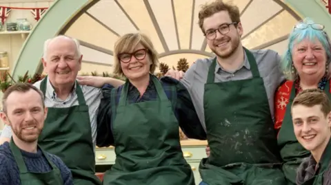 Two women and four men in green aprons, sat together in front of an arched window inside a large kitchen