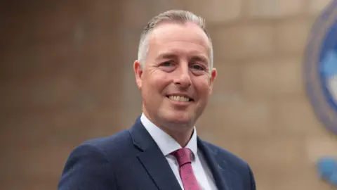 Liam McBurney/PA Wire Paul Givan smiling at the camera.  He has short, greying hair and is wearing a navy suit, a white shirt and a pink tie. 