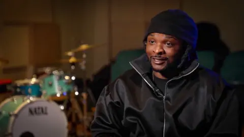 Rosca Onya in a music studio wearing black. A drum kit can be seen behind him, out of focus.