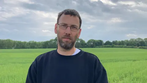 A photo showing a man with black hair and a beard. he is wearing glasses and a black jumper. The man is stood in front of a green field with tall grass, with trees in the far distance