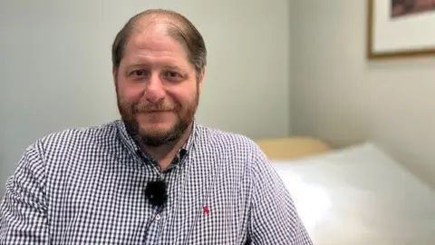 A man in a checked shirt looks at the camera. He is sitting in a plain room with little furnishing.