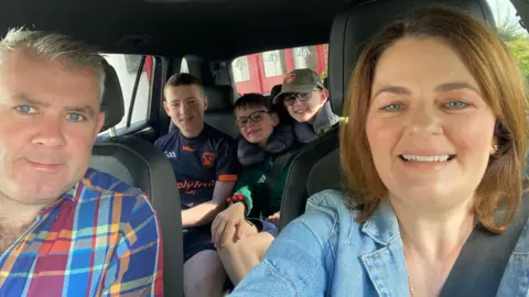 Fergal and Catherine Sherry sit in the front seat of a car while their three sons sit in the back seat. Catherine is taking a selfie and the family are all smiling at the camera. Sitting in the front passenger seat on the right, Catherine has shoulder-length brown hair and is wearing a light wash denim jacket. Fergal is sat in the driver's seat and has grey hair and is wearing a blue, red, green and yellow plaid shirt. The young boys are all bunched together in the back seat wearing dark clothing and are smiling.