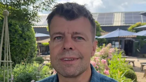 John Fairhall/BBC Chris Brown smiles at the camera. He has short dark hair and some stubble. He wears a blue shirt.