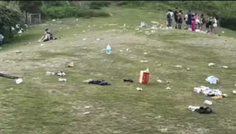 Kate Smith A large grassy area has various debris left on with a crowd of people stood in the background. Plastic bags and food packaging can be seen dotted about. 