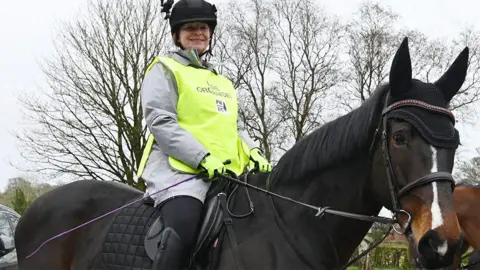 Julie-Ann Bowden in a black hard hat, grey sweatshirt and high-vis vest sits smiling on her dark brown hose with a white blaze
