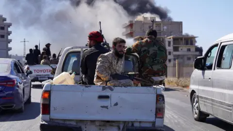 Smoke rises as Syrian security forces personnel sit in the back of a pick-up truck while entering the predominantly Druze city of Suweida, Syria (15 July 2025)