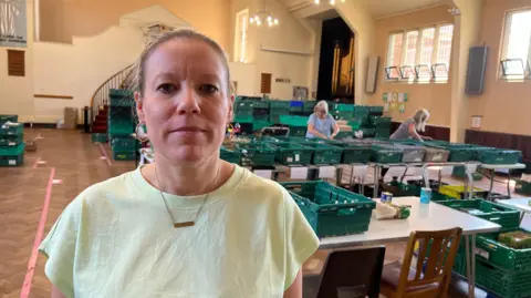 A woman is looking at the camera standing in front of rows of tables inside a large hall, with green boxes on all of them.
