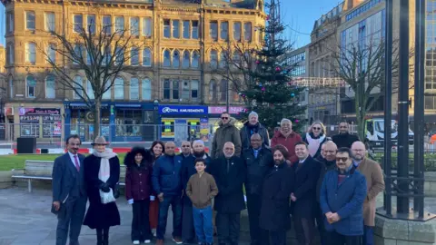 More than twenty men, women and children lined up in front of a fur tree with multi-coloured baubles hanging from it.