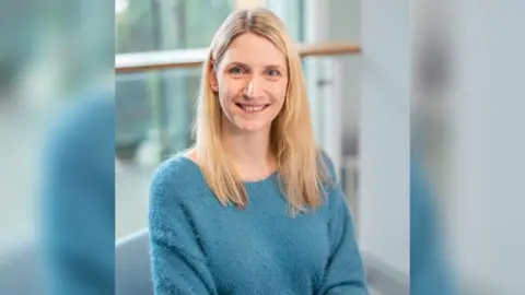 University of Hull A portrait shot of a woman with shoulder length blonde hair and a fluffy blue jumper smiling at the camera. The background is blurred and it looks like she is sat in an office-type room.
