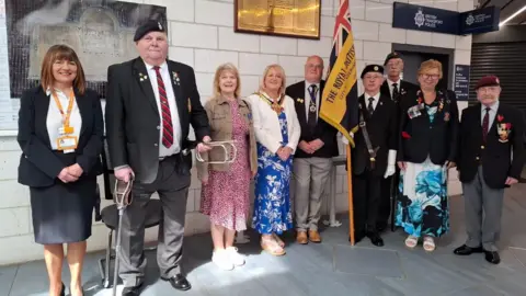 West Midlands Trains Nine people are standing up near the memorial plaque behind them. One man is carrying a flag with the Royal British Legion named on it.