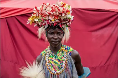 AFP A man wearing traditional clothes with a flowery head dress, colourful jewellery and a feather in his hand. He is poses for a portrait style picture.