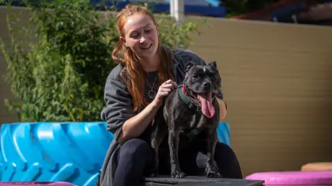 A woman with dark red hair, wearing dark clothing, pets a small black dog. The dog has its tongue out, and the woman is smiling