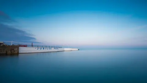 Blue and still water at Jubilee Pool in Cornwall. A white painted wall can be seen with Jubilee Pool painted on its exterior. The sky is blue.