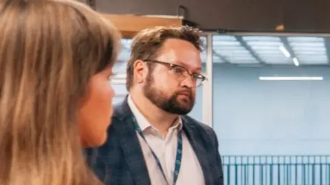 Matt Storey listens as someone speaks to him, we can also see the back of a young woman's head