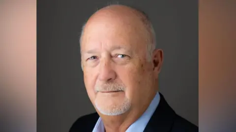 Simon Dinsdale Simon Dinsdale in a portrait-style photo. He angled slightly away from the camera but has turned his head to face the camera and smile. He wears a dark navy suit with a light blue shirt underneath. He is largely bald and has some grey facial hair.