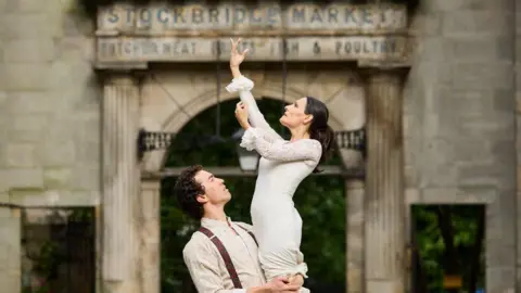 Andrew Perry A man and a woman in traditional flamenco dancing clothes. The woman is wearing a white lace dress and is being lifted by the man. She has her arm raised in the air. The man is dressed in light beige clothing with suspenders. The background features a stone archway with the words "Stockbridge Market" above it