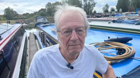 A man with grey wispy hair and a white t-shirt on, he has thin black framed glasses and is leaning on the side of a narrow boat on the water, with other narrow boats around.