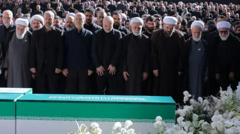Iranian President Masoud Pezeshkian, Iran's Parliament speaker Mohammad Baqer Qalibaf and Chief Justice of Iran Gholam-hossein Mohseni Ejei with officials attend a prayer during a public farewell ceremony to pay their respects to late Supreme Leader Ayatollah Ali Khamenei. A large crowd can be seen in the background as everyone stands looking at the coffin