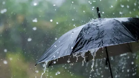 A black umbrella is covered with falling water that looks like rain. The background is blurred. 