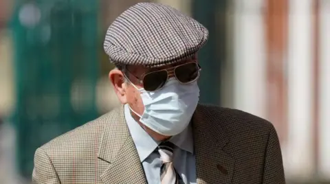 PA Media A man in a tweed cap is wearing dark glasses and a mask, which covers much of his face. He also has on a tweed jacket, a grey shirt and a striped tie.