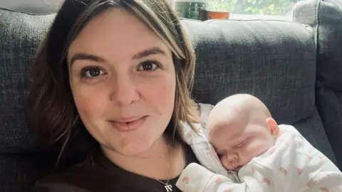 New mum Chloe with shoulder length brown hair wearing a black t-shirt with her sleeping baby wearing a white and pink sleep suit resting on her shoulder. Chloe is sitting on a grey sofa.