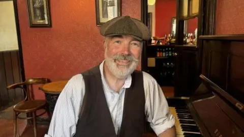 A man in a flat cap, white shirt and waistcoat sitting in a room with red walls beside a piano