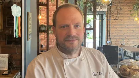 Grace Wood/BBC A man with short light brown hair stands in a cafe. He is wearing chef whites