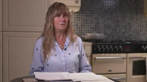 Tracy Heaton has long blonde hair and is wearing a blue and white striped shirt. She is sitting at her kitchen table, with letters laid out in front of her.