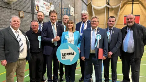 A group of nine men and one woman are standing in a sports hall. They are mostly wearing suits. The woman is standing in the centre and is holding a light blue Reform UK sign.
