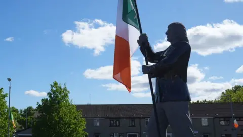 A metal statue of a long haired Bobby Sands, holding a green white and orange Irish flag, there is a row of brown, residential terraced houses in the bakcground and some large green trees