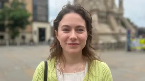 Eva Martin is young with long brown hair and blue eyes. She is wearing a light green cardigan and white top.