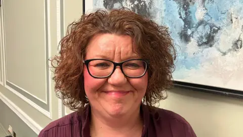 Julie Graham has short brown curly hair and is wearing makeup, black glasses and a purple top.