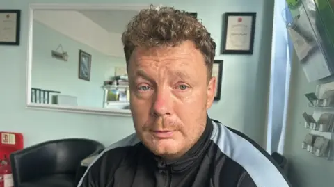 Jason Burrows seated indoors wearing a black zip-up sports top with light grey sleeves. The setting includes pale blue walls, framed certificates, a wall mirror, a black armchair, and brochure holders mounted on the wall beside a window.