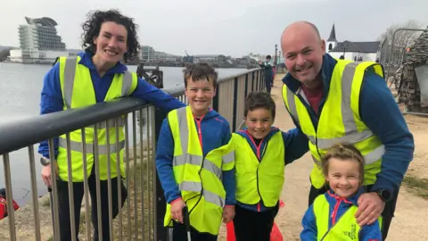 The Power family smile at the camera. A mum stands behind the railings, she is wearing a blue jumper, high vis yellow jacket, three children (two boys and a little girl) stand between her and her partner. All are wearing blue jumpers and high vis yellow vests. The Norweigan church in Cardiff is visible behind them.