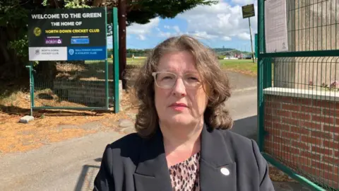 Kellie Armstrong, a woman with shoulder-length brown hair, looks into the camera outside of the cricket ground. She is wearing grey-rimmed glasses and a brown and white top. In the background there are green metal fences, trees and grass.