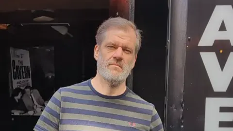 A wide shot of David Dutton standing outside the Cavern Club. He has grey hair and is wearing a stripy t-shirt.