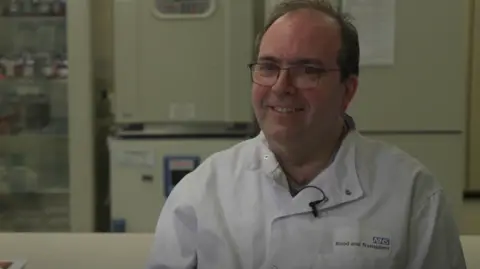 Toni Woodward wearing his working overalls wearing glasses and smiling at the camera. He is sitting in a lab.