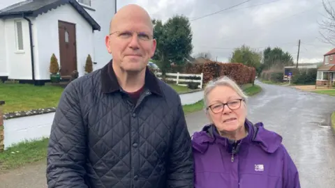 A man with a black jacket on who is wearing glasses and a shorter woman next to him with blonde hair tied back, glasses and a purple coat are smiling into the camera. They are stood down a lane next to a few houses in a countryside village.